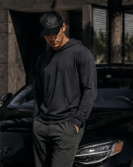 Person wearing a black hoodie and golf cap standing next to a black car.