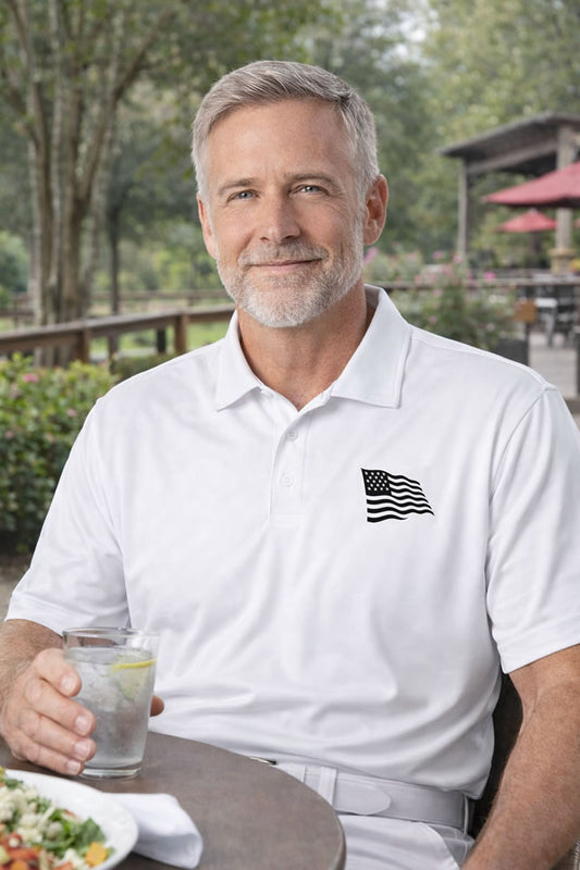 Man in a white polo shirt with an American flag emblem, holding a drink outdoors.